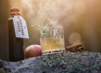 I motljus står Swedish Tonic Christmas spices tonic. på en vacker sten. Till höger i bild, en drink och glöggkryddan kanel, här i form av två kanelstänger.