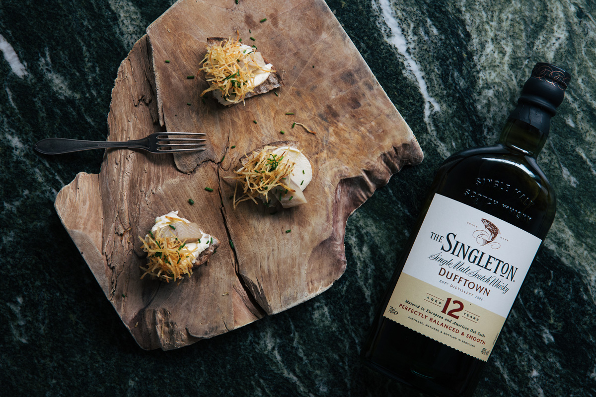 Några läckra matbitar på en bohemisk träskiva, en ensam gaffel och en flaska Singleton whisky fotograferat på Tjoget av Petter Bäckström